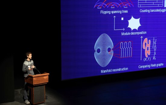 A man speaks from a podium next to a projection of mathmatical images.
