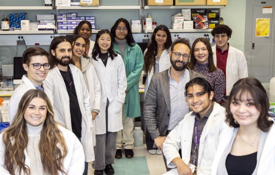 Image shows 13 smiling people, including a mix of genders, in a science lab. Most are students wearing white lab coats. Shelves on the back wall hold boxes of lab supplies.