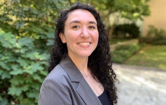 A young woman with dark curly hair wearing a gray blazer smiles for a portrait. Bushes and greenery are blurred in the background.