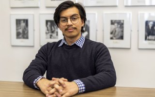A young man with dark hair and round glasses, wearing a charcoal sweater, sits at a wooden desk with his hands folded, looking at the camera. The wall behind him is white and is hung with a series of black and white photos in equal-sized silver frames.