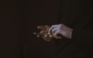 A pair of hands can be seen holding a phone in a dark room. The lit screen is the only source of light.
