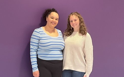 Two young women, one in a striped blue sweater and one in a beige sweater, stand smiling in front of a purple wall.