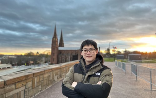 A man with black hair and glasses stands with his arms folded against a gray setting sky with a cathedral rising in the background.