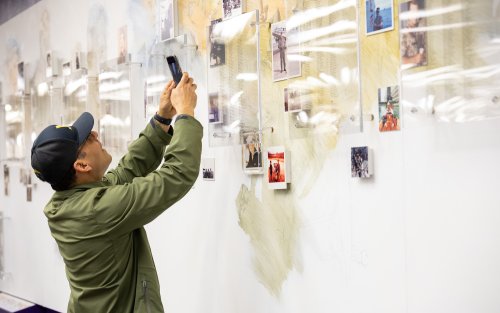 A man in a green jacket and dark cap takes a photo of a wall adorned with photos and artist renderings of UAlbany veterans