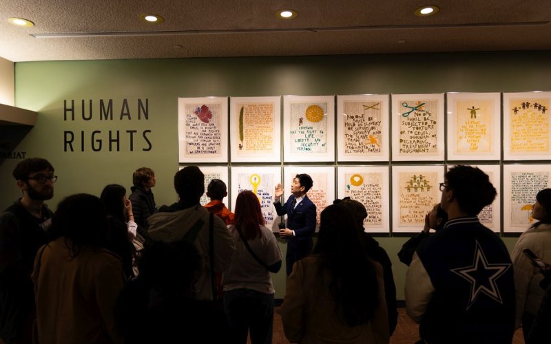 The image features a wall where 14 paintings are hung in two even rows of seven. To the left on the wall are the words ‘Human Rights’. A man wearing a blazer gestures to the paintings. A group of about a dozen students can be seen, in silhouette, in the foreground.  
